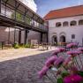 Ein Innenhof mit Kopfsteinpflaster, Tischen und Stühlen, umgeben von Gebäuden mit Arkaden und einem modernen Balkon.