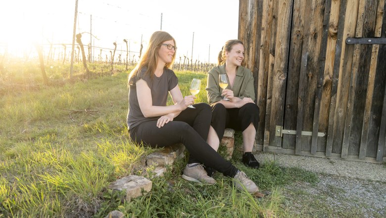 Zwei Frauen sitzen mit Weingläsern vor einer Holzhütte im Weinberg.