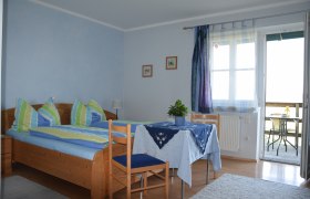 Gem&uuml;tliches G&auml;stezimmer mit Doppelbett, Tisch und St&uuml;hlen, blauen Vorh&auml;ngen und Zugang zu einem Balkon mit Sitzgelegenheit.